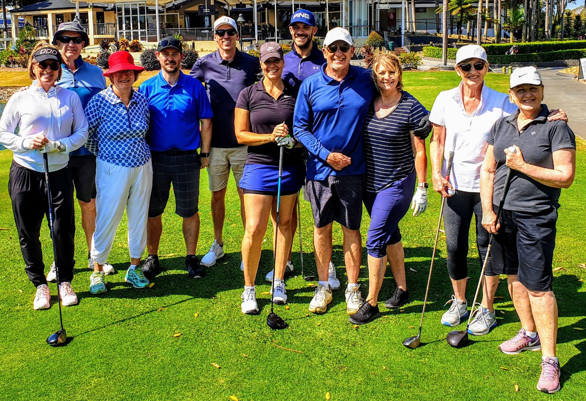 Group of men and women posing with golf clubs on a sunny day at a golf course with a clubhouse in the background. . GOLF COACHING, PAUL WILLIAMSON GOLF