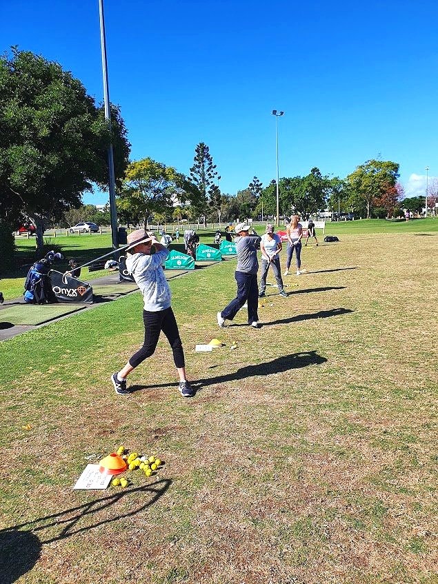 GOLF LESSONS, GOLF COACHING, GOLD COAST GOLF, EMERALD LAKES GOLF CLUB. People practicing golf at a driving range, with several individuals swinging clubs on a grassy area. Golf bags are seen in the background under a clear blue sky.