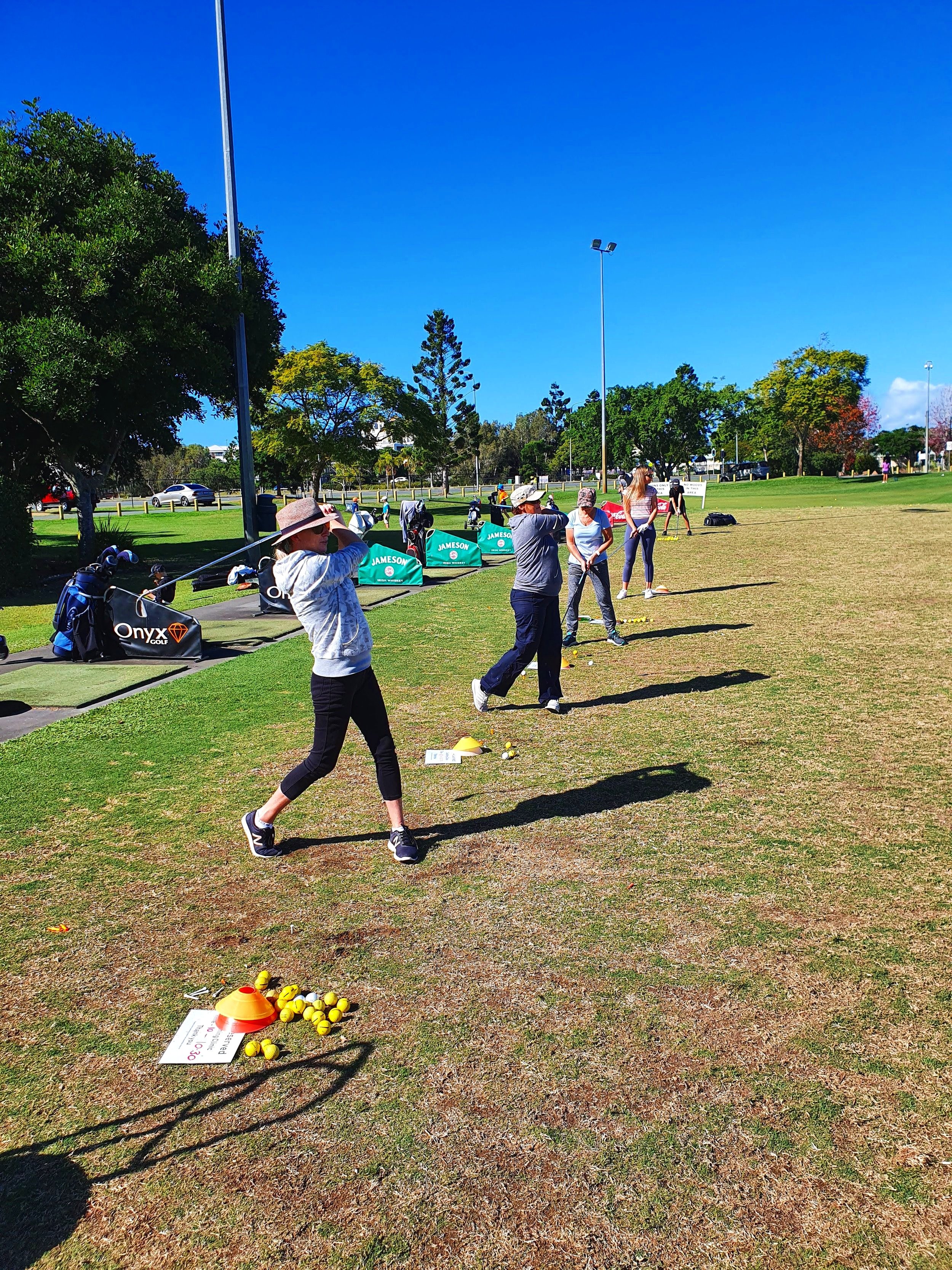 People practicing golf swings on a driving range under clear blue sky, with golf equipment and trees in the background.