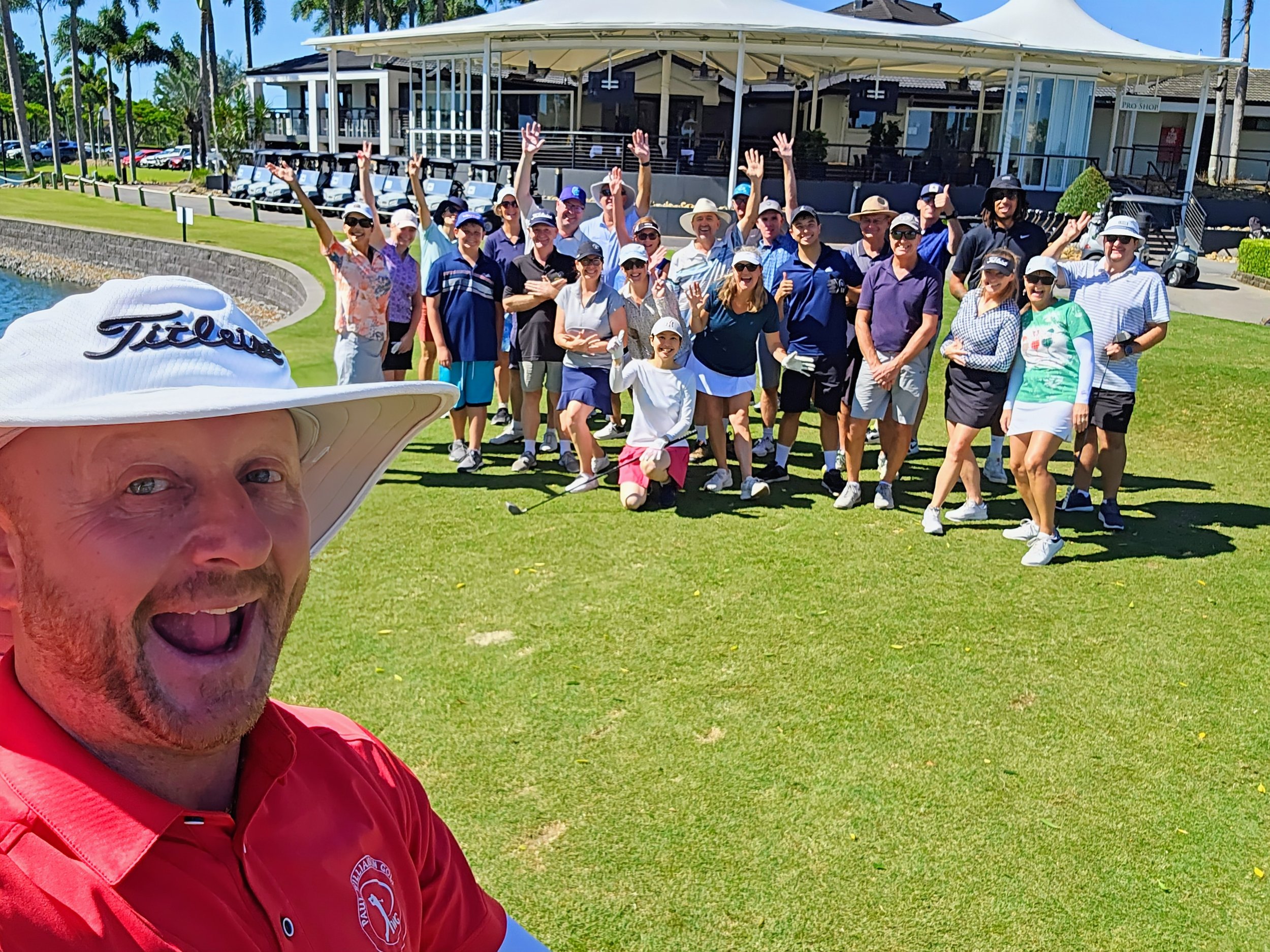 Group of people posing for a selfie on a golf course, with clubhouse and palm trees in the background.