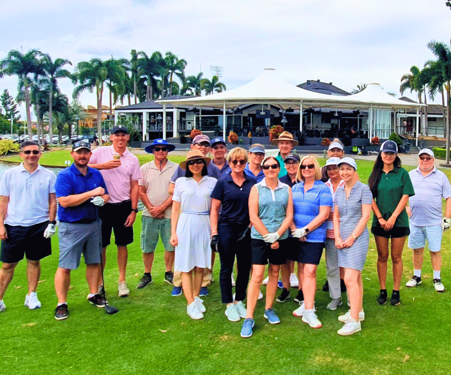 Group of people posing on a golf course with clubhouse in background, wearing casual and golf attire, palm trees nearby.