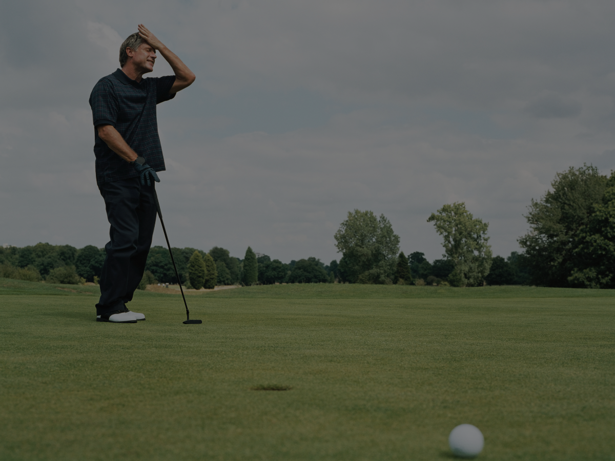 Person on golf course looking disappointed after missing putt, with golf club in hand, golf ball near hole, and trees in background.