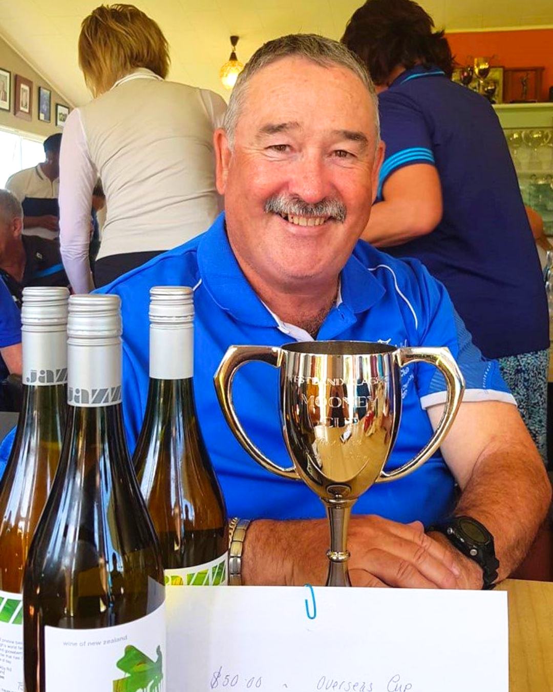 A smiling man in a blue sports shirt sitting at a table with four bottles of wine and a shiny gold trophy cup, celebrating at a gathering.