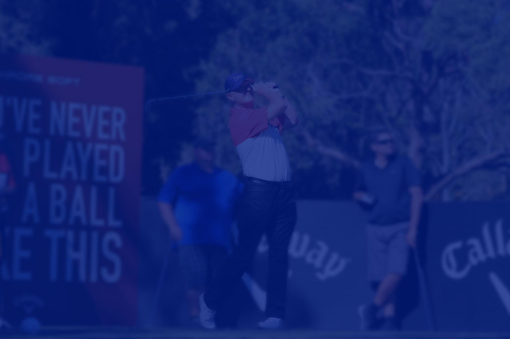 Golfer swinging club on course with spectators and signage in the background.