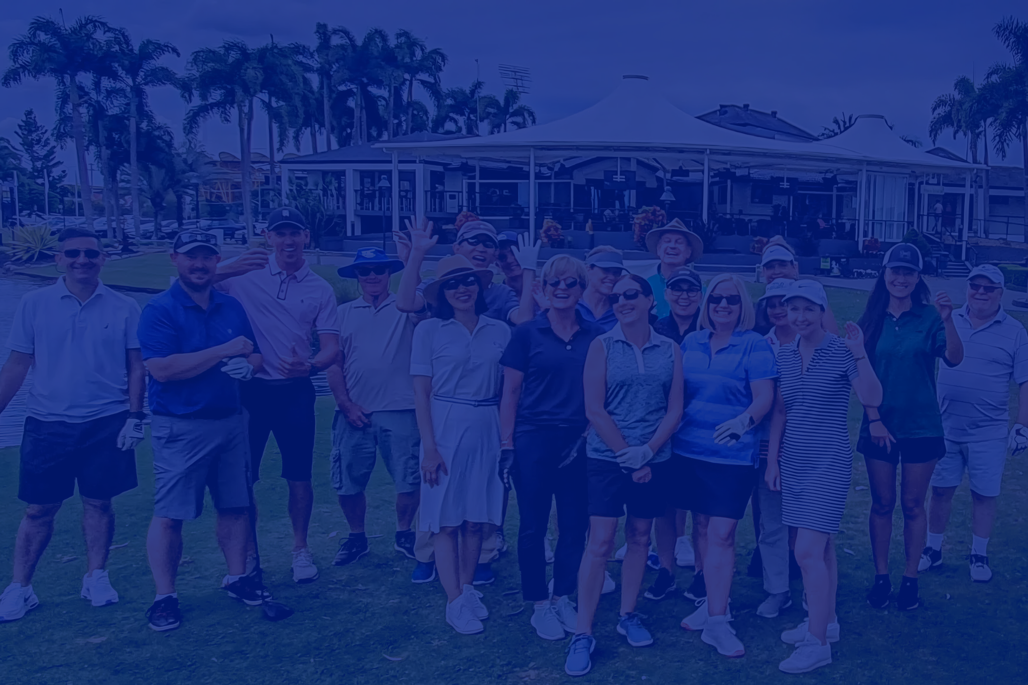 Group of people posing at a golf course, wearing casual golf attire, with a clubhouse and palm trees in the background.